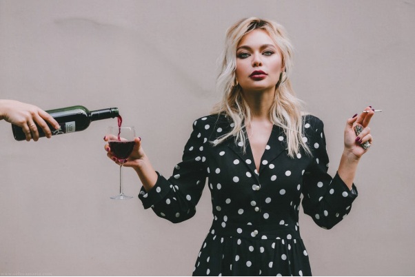 A Woman in a Black Polka Dotted Dress Holding Out Her Wineglass to be Filled on the Right and Clutching a Cigarette and Lighter in her Left Hand