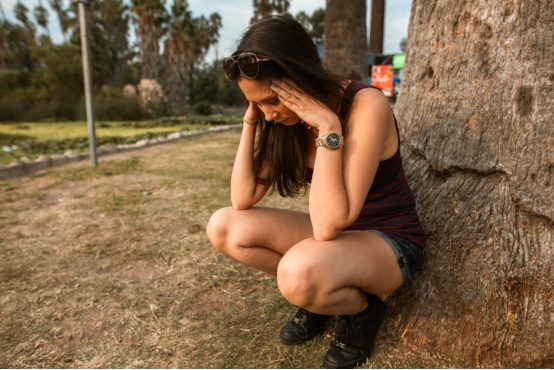 A woman sits back against a tree while holding her head, looking anxious and stressed