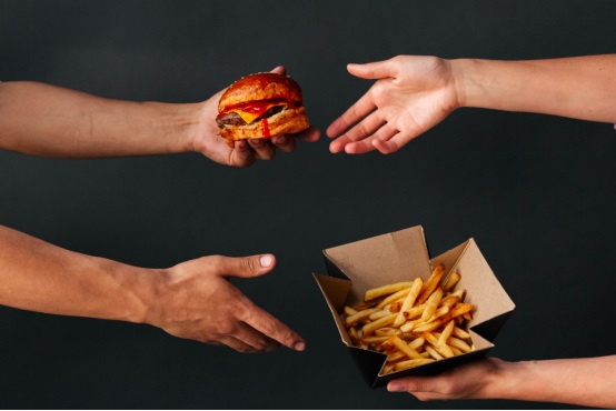 Two sets of hands exchanging a burger and fries, both unhealthy fast foods