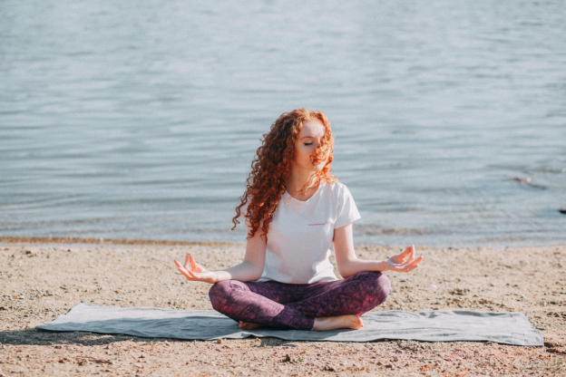 A woman sitting near the ocean meditating on a mat